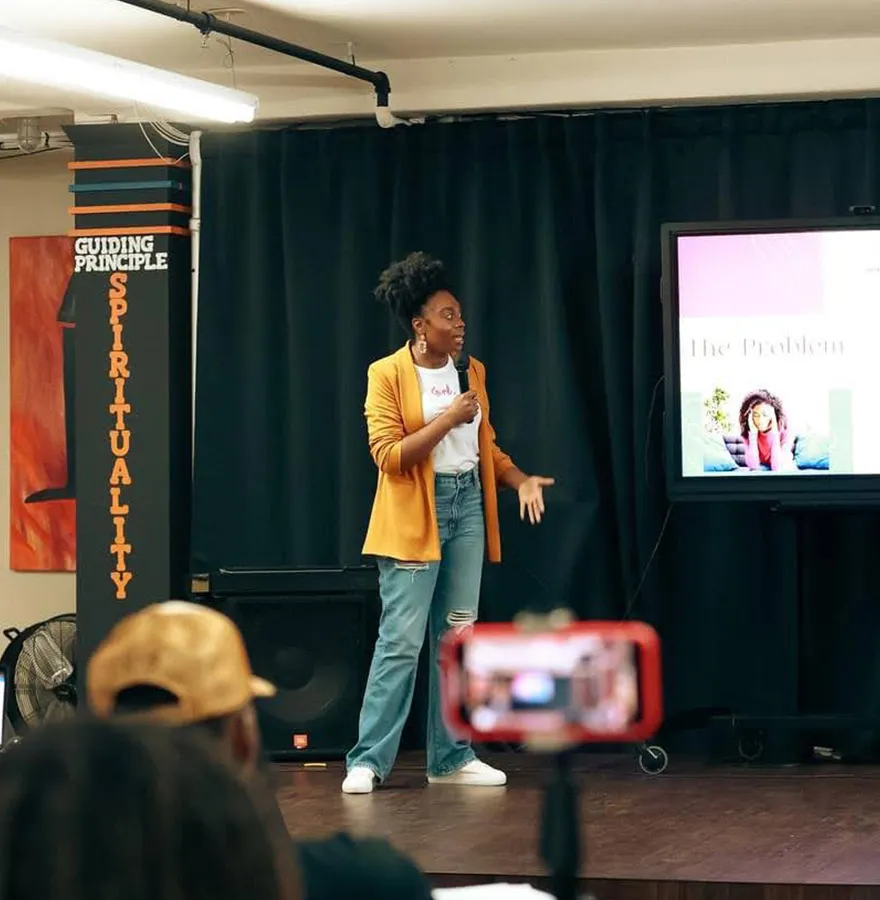 An African American woman in a yellow blazer speaks on stage with a microphone, gesturing toward a screen displaying a presentation.
