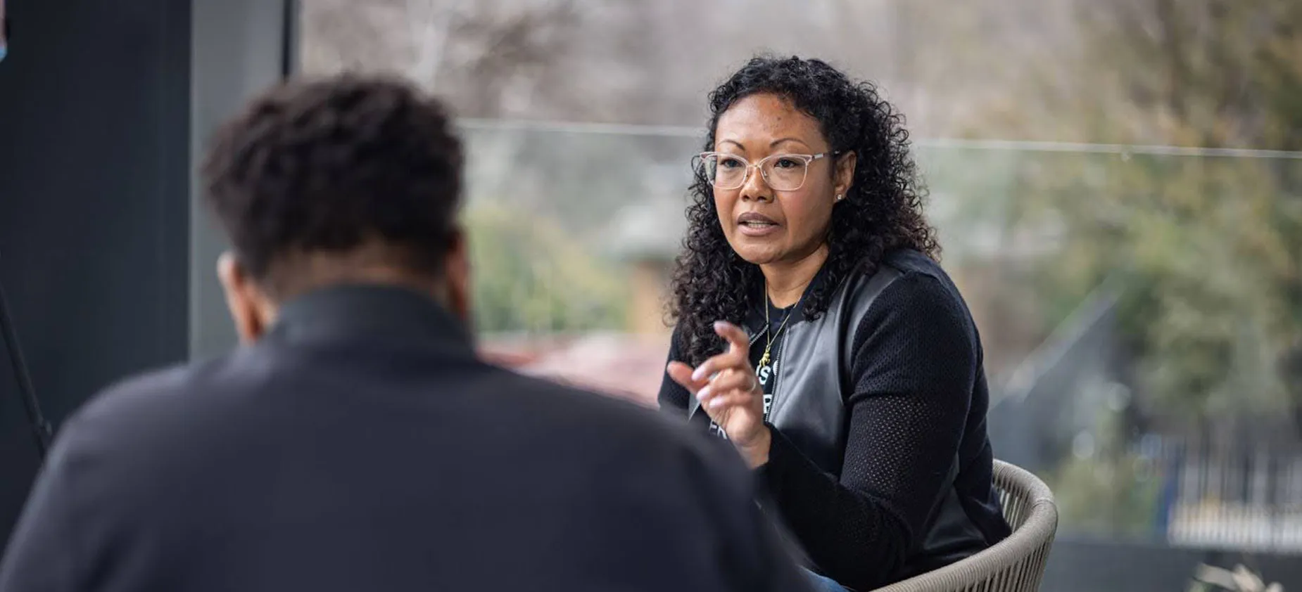 A middle aged African American woman with glasses sits in a chair talking to another person whose back is to the camera.