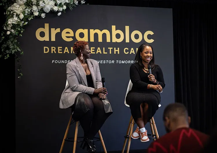 Two women sit onstage, speaking at a "dreambloc DREAM WEALTH CAMP" event. One holds a microphone, and both are smiling.