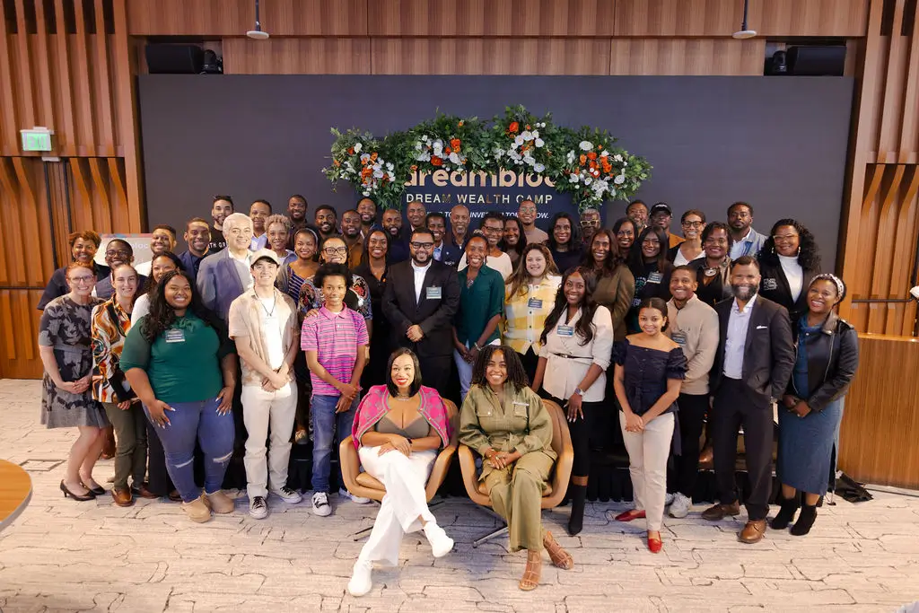 A large group of people pose together indoors at an event, standing and smiling, with a decorated backdrop and a mix of formal and casual attire.